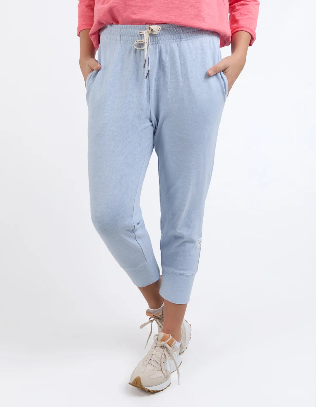 Image of A woman wearing Elm light blue cropped jogger pants with a drawstring waist, paired with a coral pink top and beige sneakers, shown against a plain white studio background.