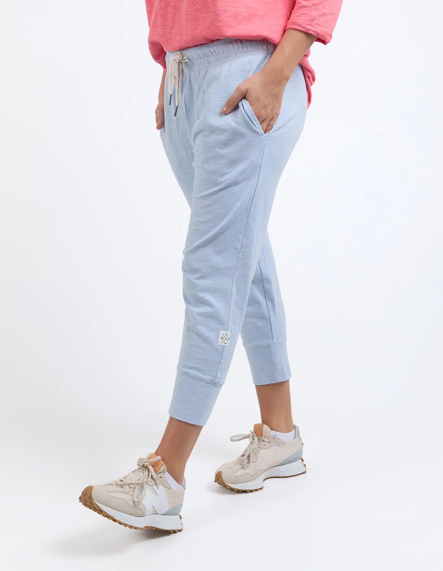 Image of A woman wearing Elm light blue cropped jogger pants with side pockets, a pink top, and beige sneakers, shown from the waist down against a plain white studio background.