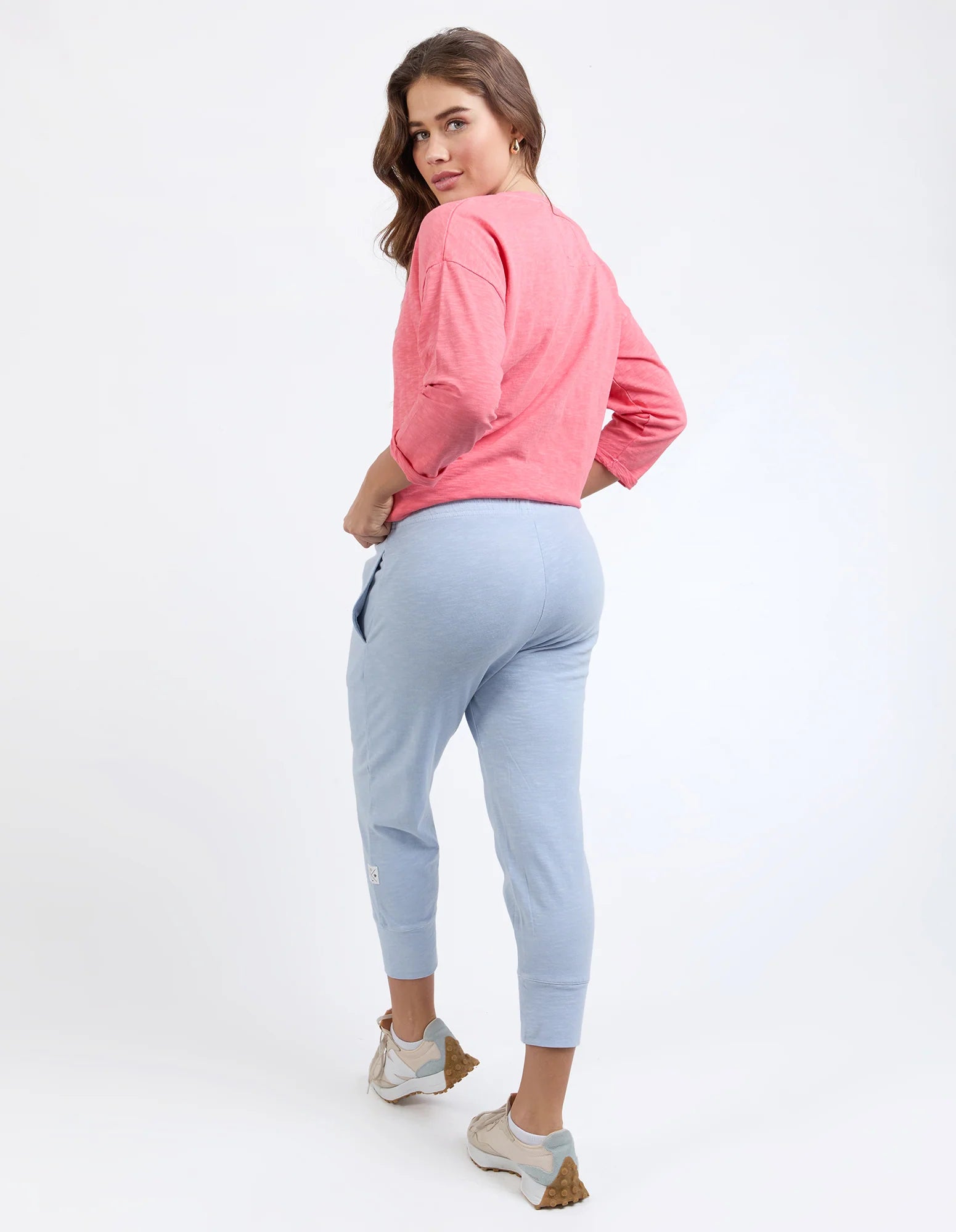 Image of A woman modeling Elm light blue jogger pants and a pink long-sleeve top, shown from a back-three-quarter angle against a plain white studio background.