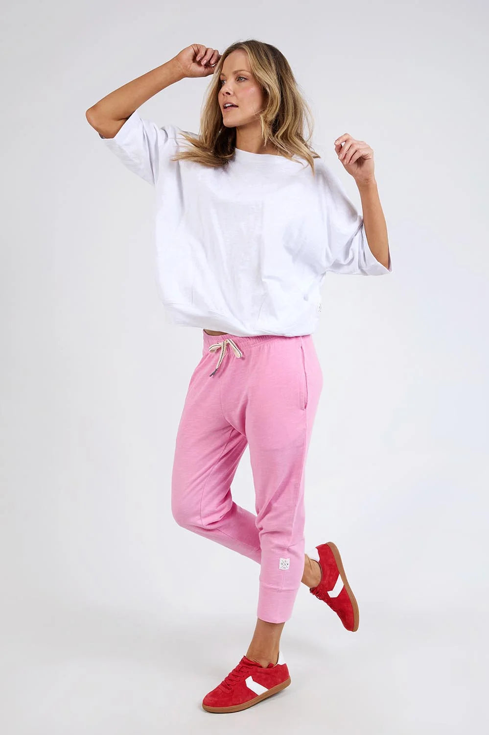Image of A woman modeling a white oversized short-sleeve top, Elm pink jogger pants, and red sneakers against a light gray studio background.