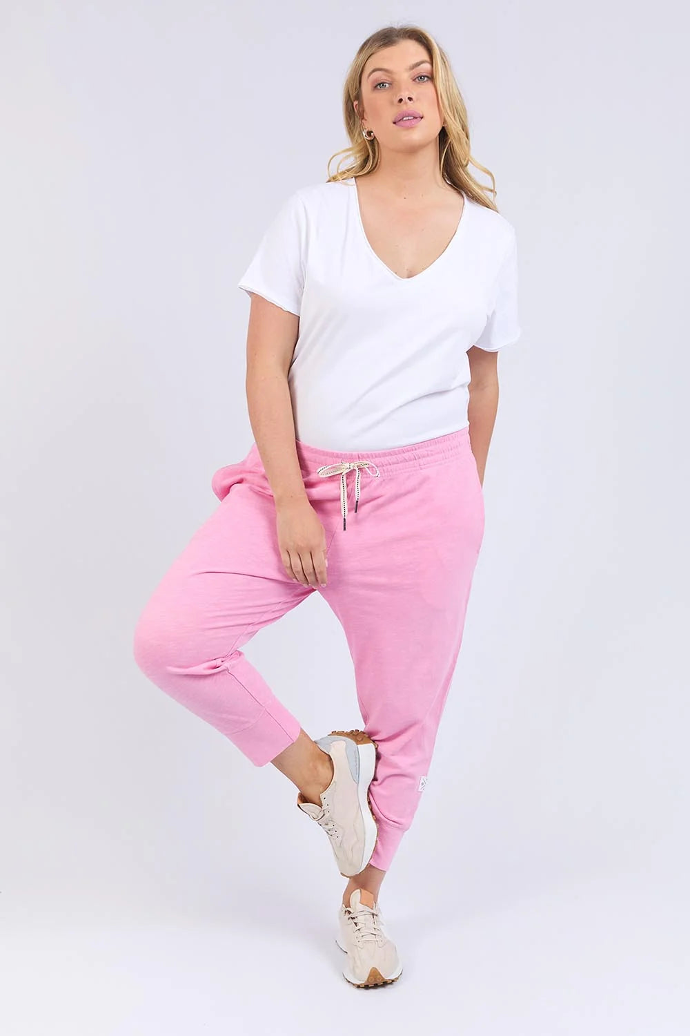 Image of A woman modeling Elm pink jogger sweatpants with a white V-neck t-shirt and white sneakers, standing against a light gray studio background.