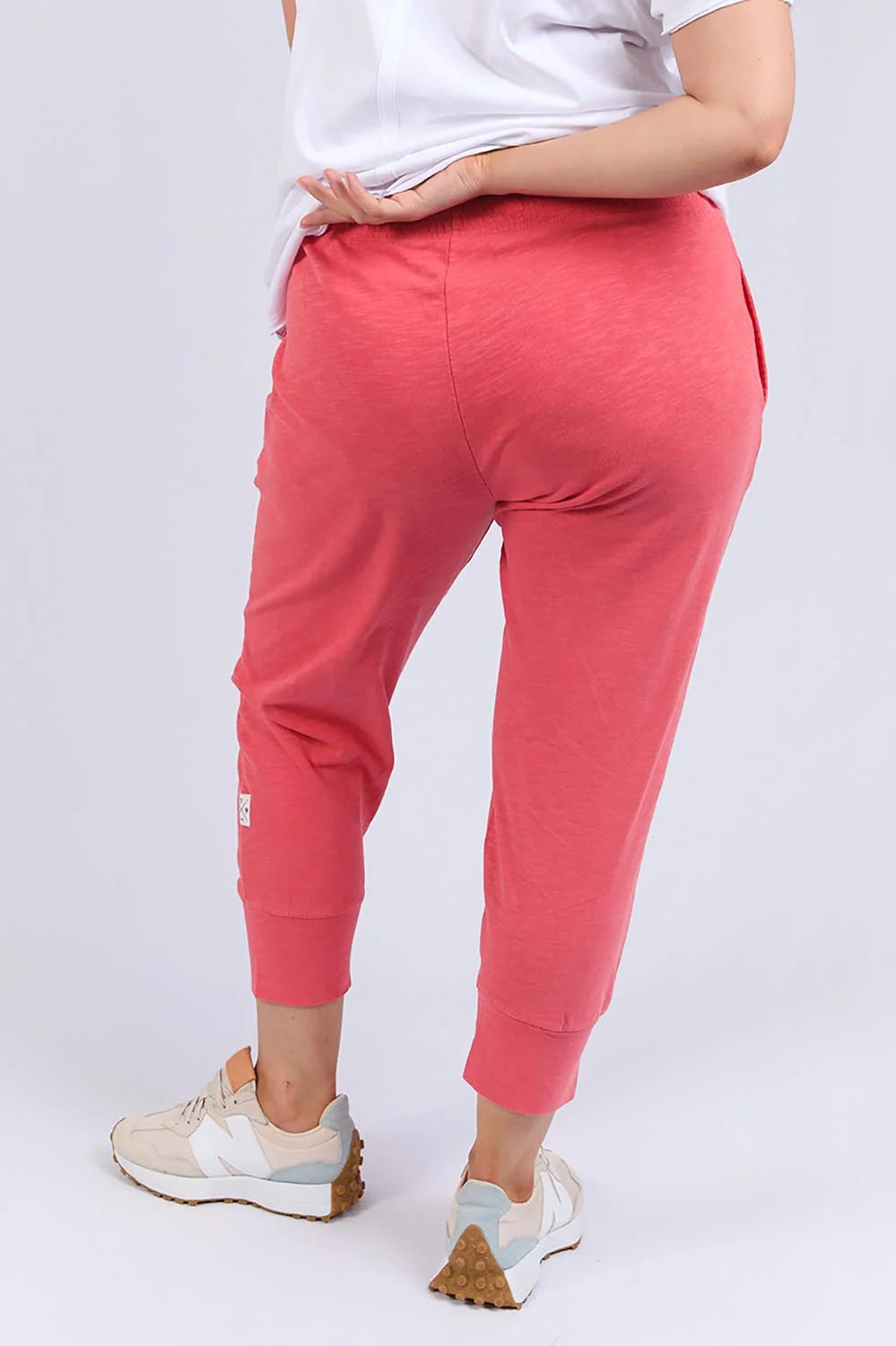 Image of A woman shown from the back wearing Elm bright coral pink jogger-style pants with a white top and white sneakers, photographed against a light gray studio background.