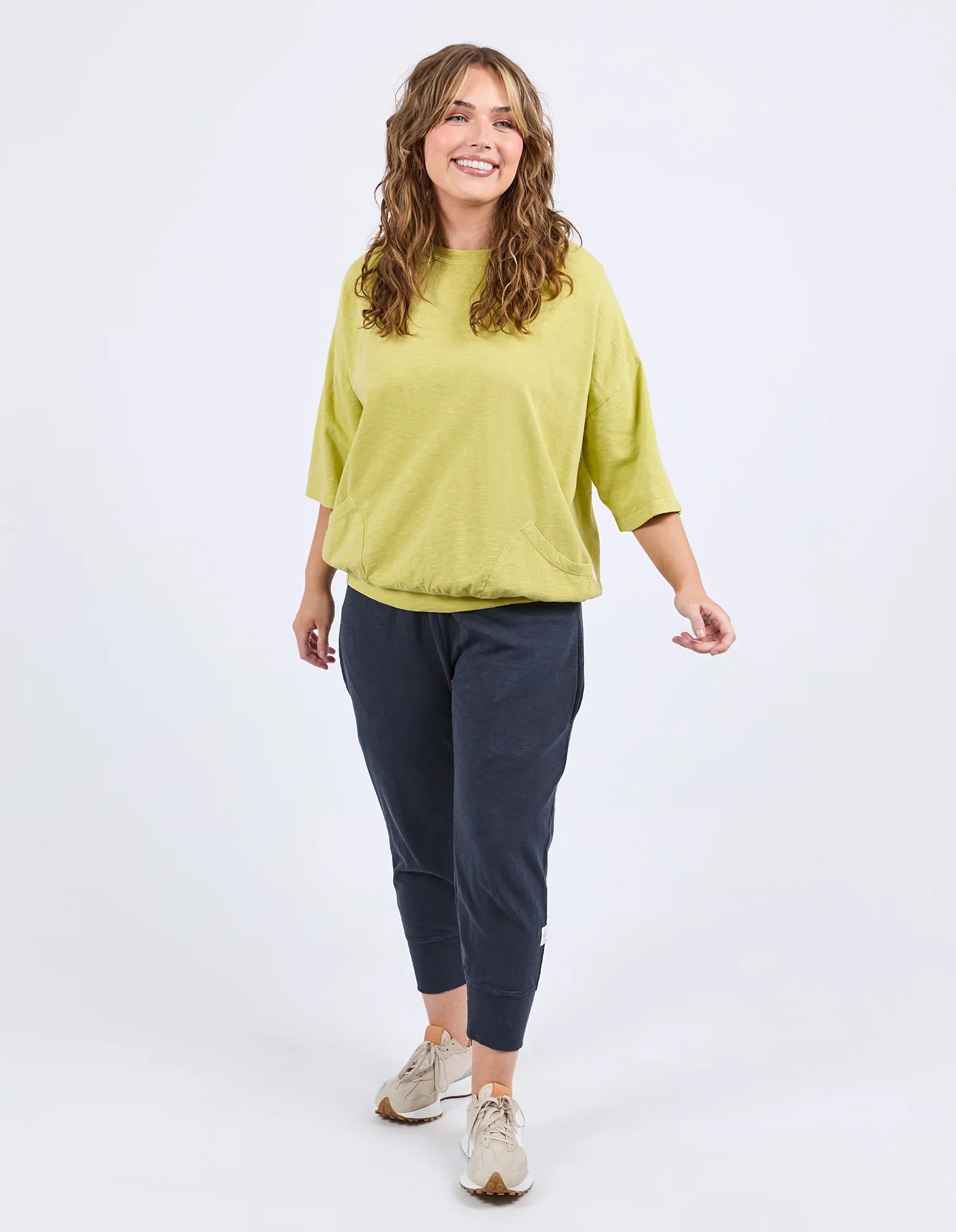 A woman modeling a loose-fit Elm lime green short-sleeve top and dark navy cropped pants with beige sneakers, photographed against a clean white studio background.