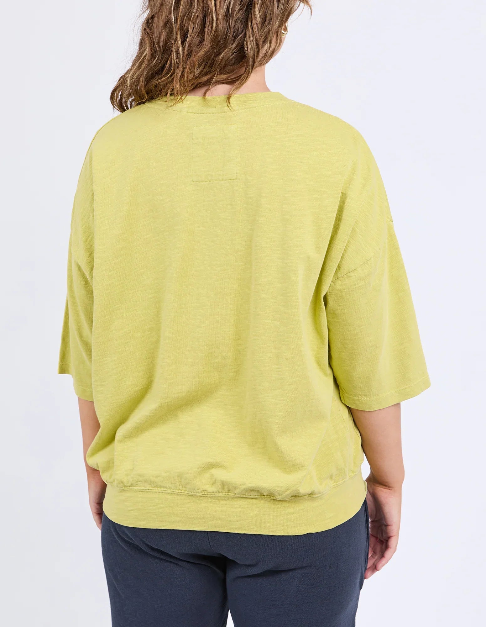 Image of A woman viewed from the back wearing a loose, Elm short-sleeve lime green top and dark navy pants against a plain white background.