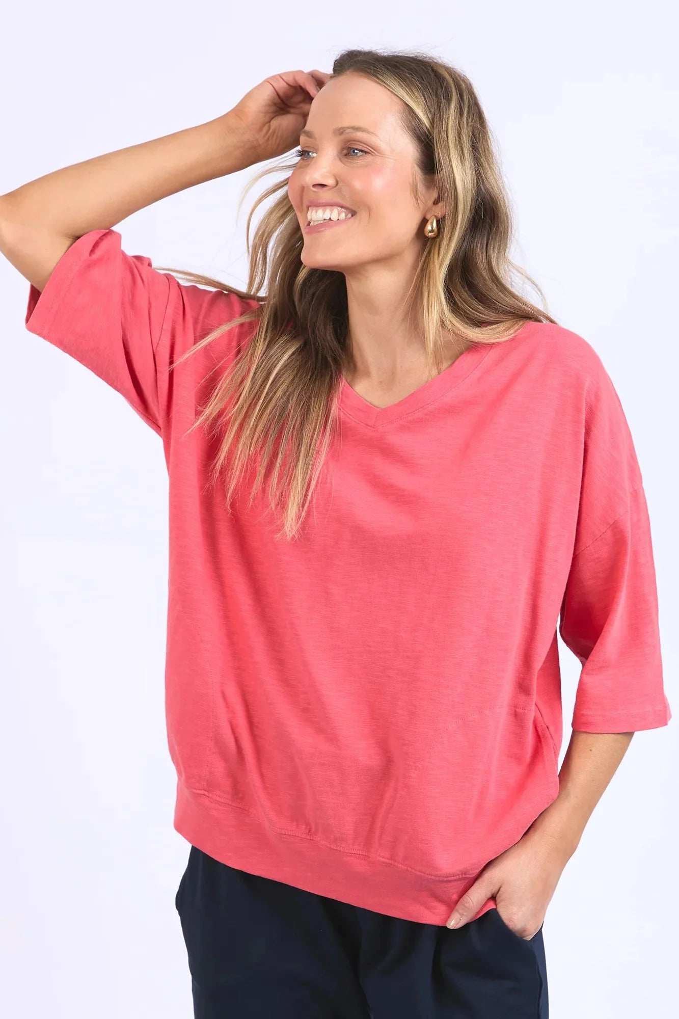 Image of A woman wearing a Elm coral pink V-neck short-sleeve top and dark pants, posing against a light gray studio background.