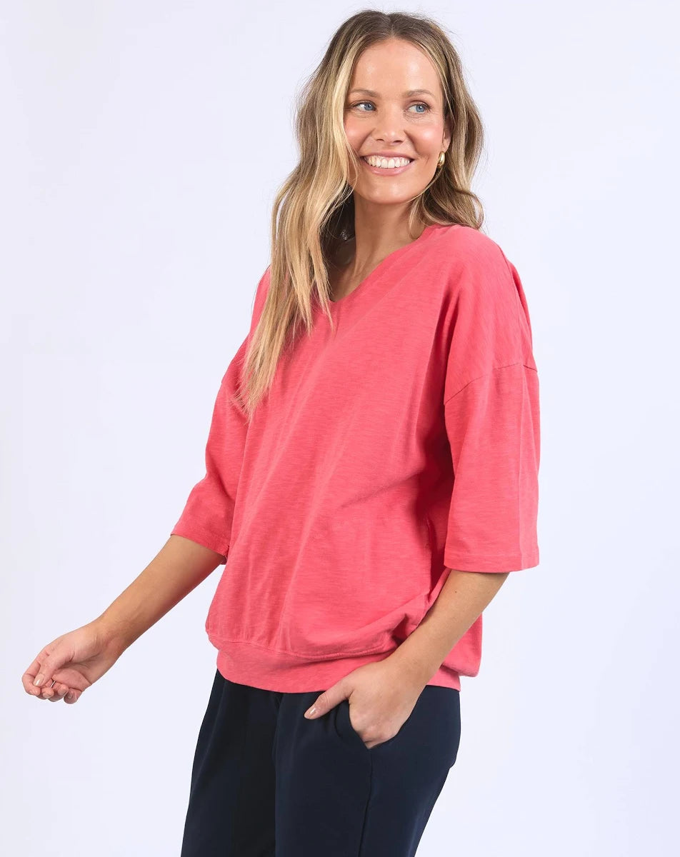 Image of A woman modeling a Elm coral pink V-neck knit top with three-quarter sleeves and dark navy pants, standing against a light gray studio background.