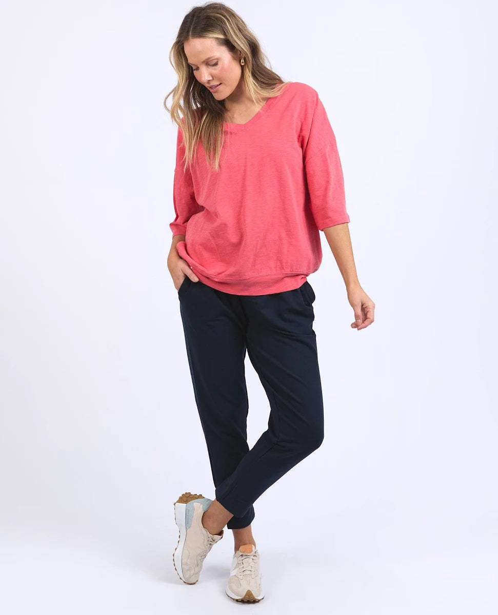 Image of A woman modeling a Elm coral pink three-quarter sleeve top and dark navy pants with beige sneakers, standing against a plain white studio background.
