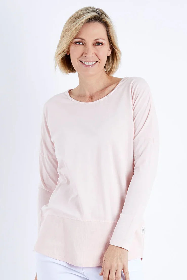 A smiling blonde woman wearing a Elm light pink long-sleeve crewneck top and white pants, photographed against a clean white studio background.