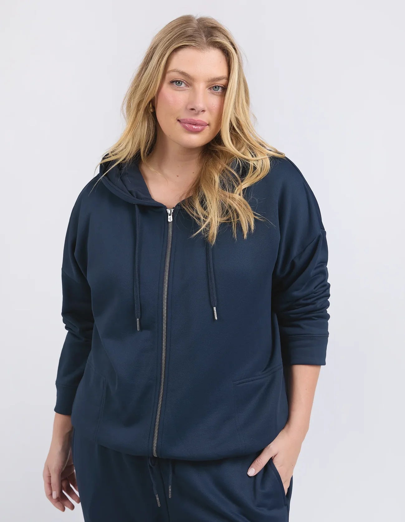 Image of A woman modeling a Elm navy blue zip-up hoodie and matching sweatpants against a light gray studio background.
