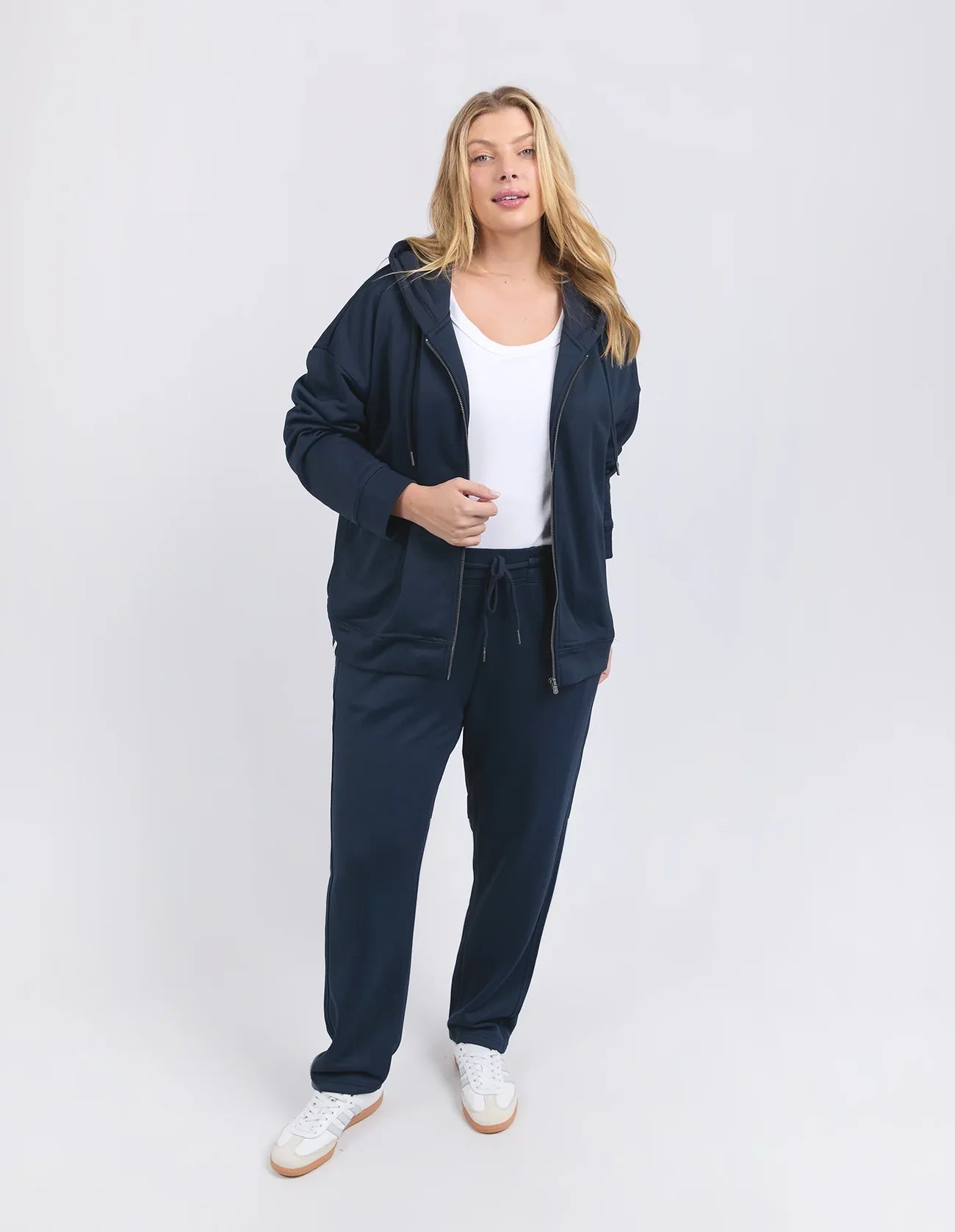 Image of A woman modeling a Elm matching navy blue lounge set consisting of a zip-up hoodie and sweatpants, worn over a white tank top and white sneakers, against a light gray studio background.