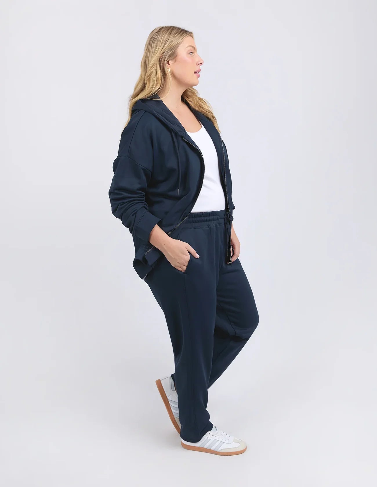 Image of A woman modeling a matching Elm navy blue lounge set consisting of a zip-up hoodie and sweatpants, styled with a white tank top and white sneakers, against a light gray studio background.