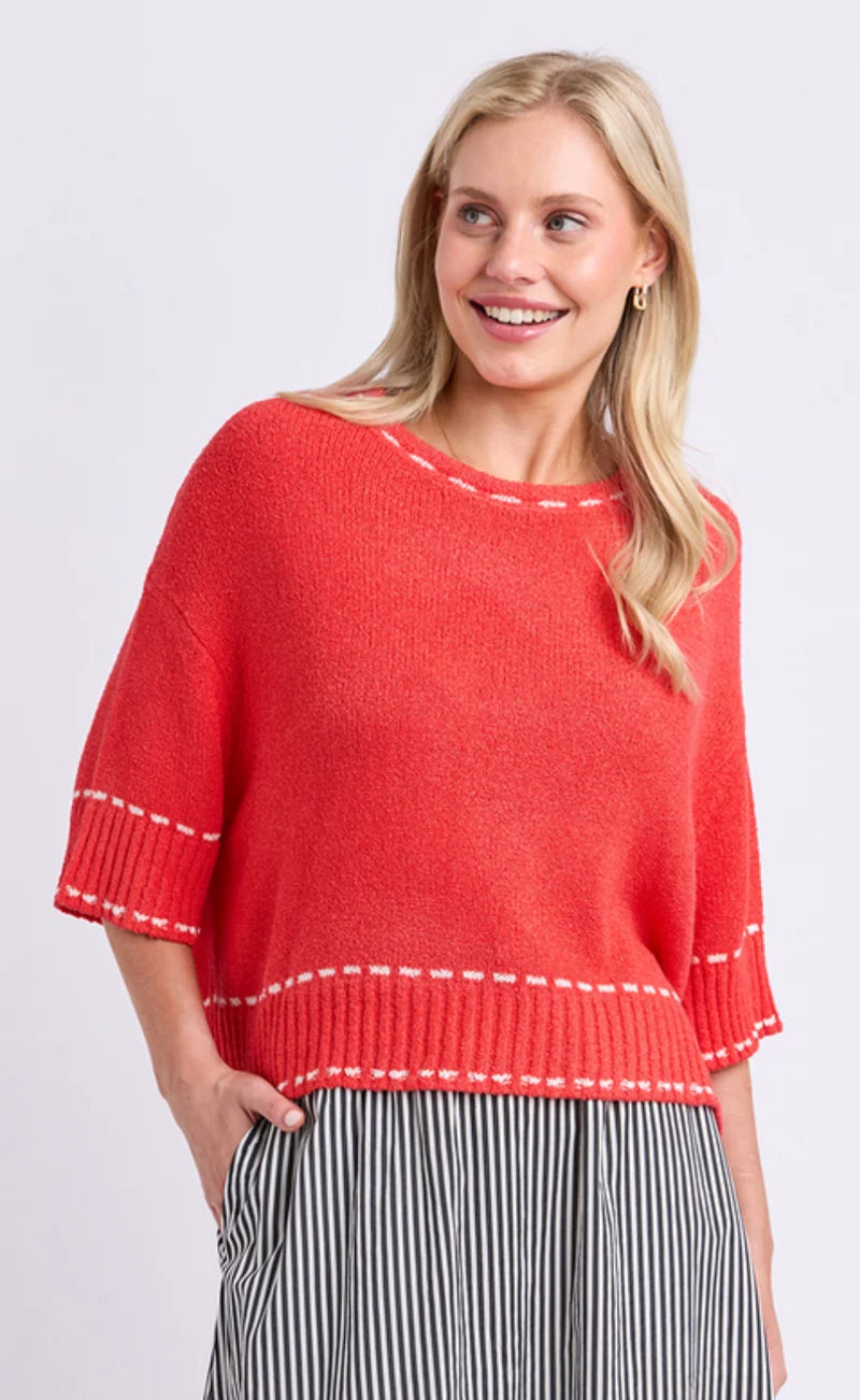 Image of A smiling blonde woman wearing a Elm bright coral-red knit sweater with white contrast stitching and striped black-and-white pants, photographed against a light gray studio background.