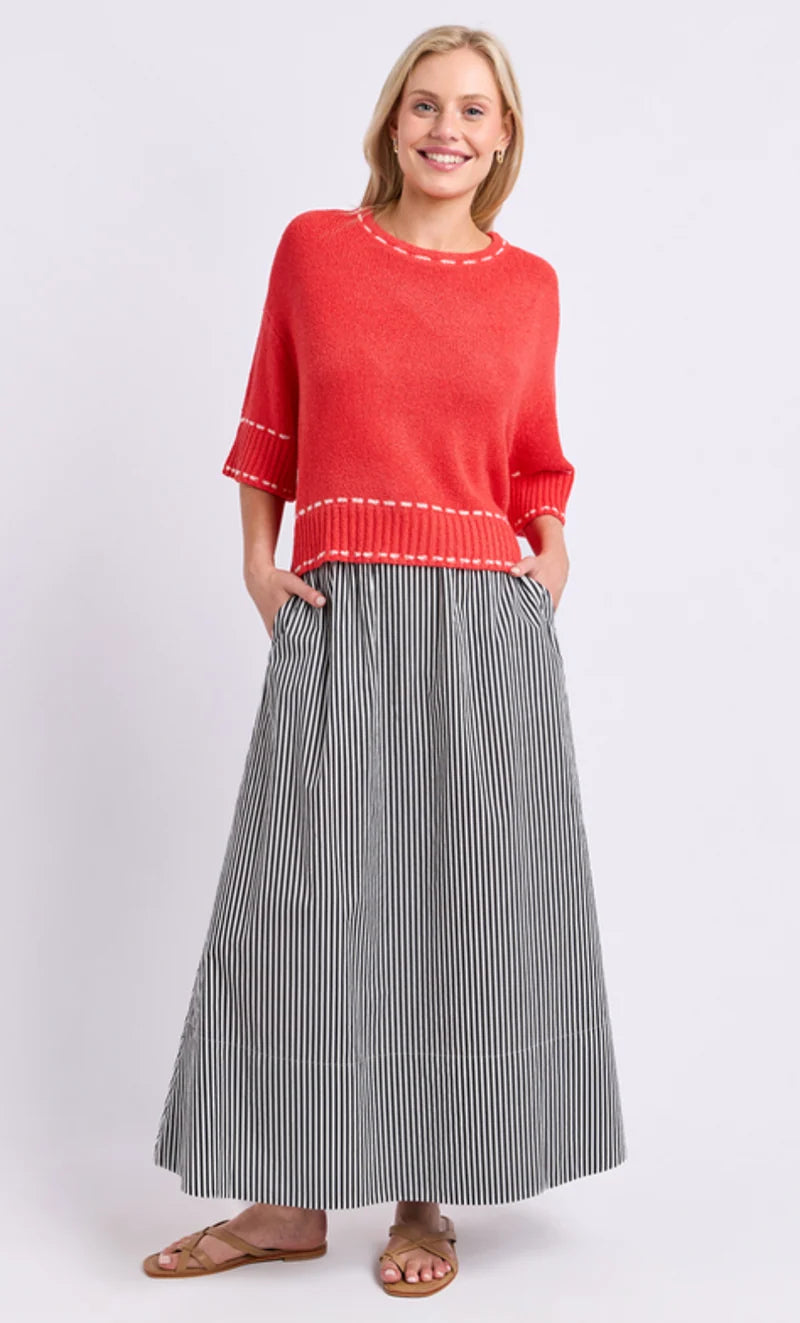 Image of A woman modeling a Elm bright coral-red knit sweater with white contrast stitching and a long gray-and-white vertically striped skirt, paired with brown sandals, against a light gray studio background.