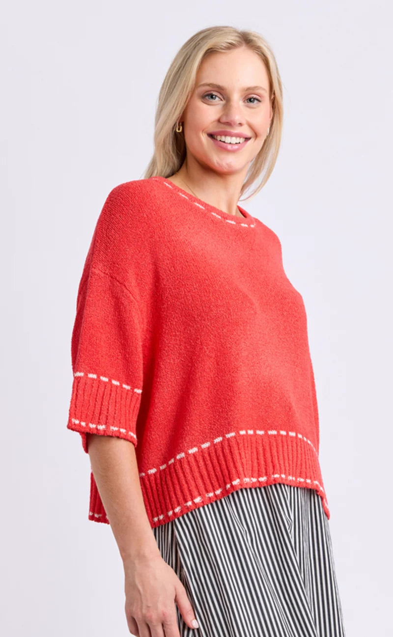Image of A smiling blonde woman wearing a Elm coral-red knit sweater with white contrast stitching and striped pants, photographed against a light gray studio background.