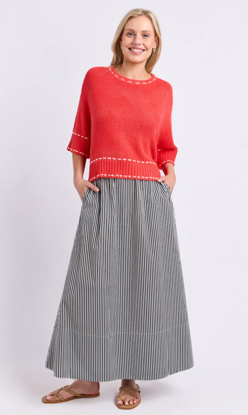 Image of A woman modeling a Elm bright coral-red knit sweater with white contrast stitching and a gray-and-white vertically striped midi skirt, paired with brown flat sandals, against a light gray studio background.