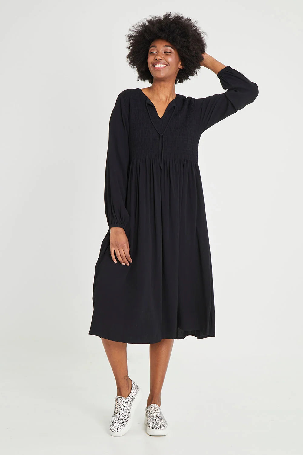 A woman modeling a Elm black long-sleeve midi dress with a V-neckline and gathered waist, paired with silver sneakers, against a plain light gray studio background.