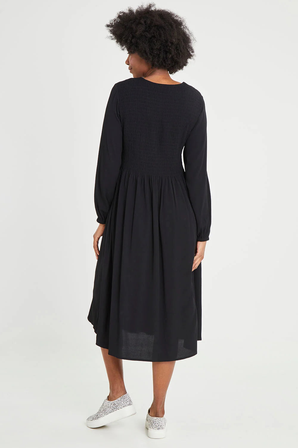 Image of A woman with curly black hair is shown from the back wearing a Elm long-sleeved black midi dress and sparkly silver slip-on sneakers against a plain white studio background.
