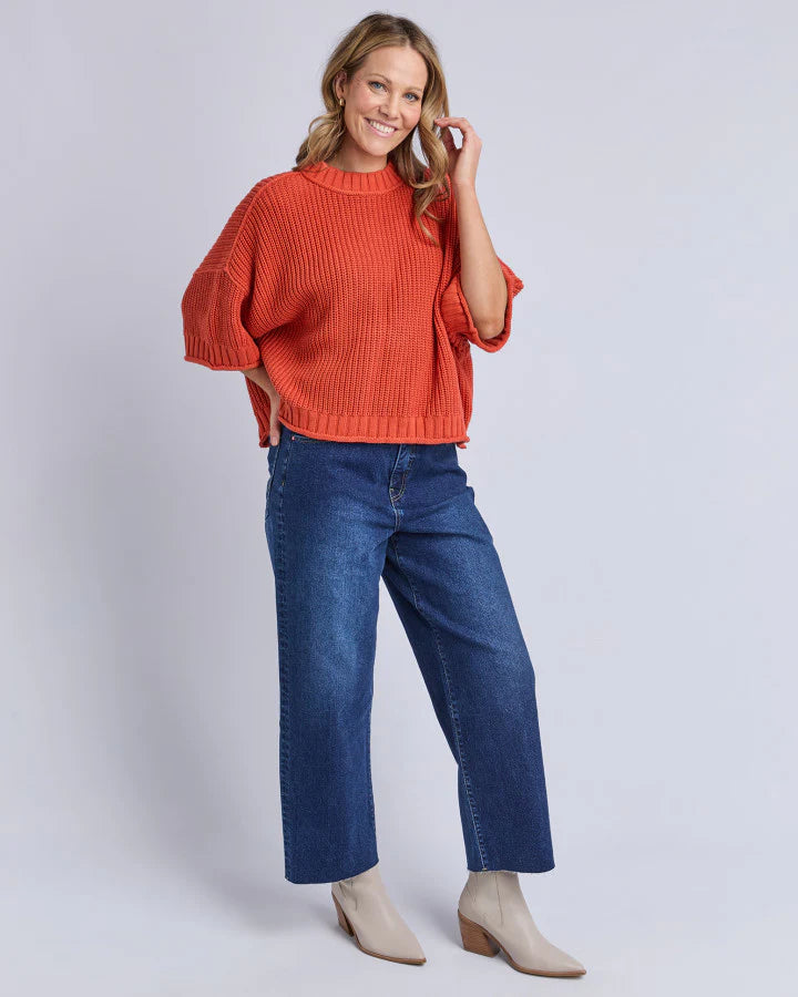 Woman wearing an Elm orange sweater and blue jeans on a white background