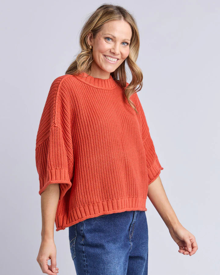 Woman wearing a Elm rust-colored sweater and blue jeans on a light gray background
