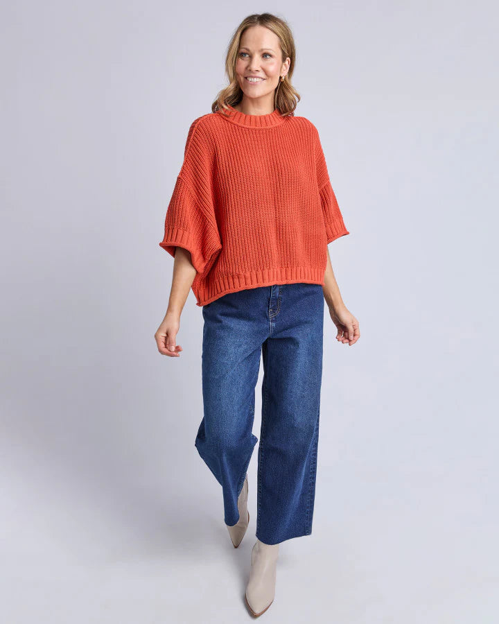 Image of Woman wearing an Elm orange knit and blue jeans on a light gray background