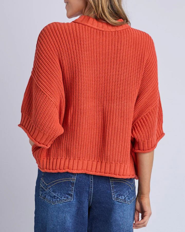 Image of Woman wearing an Elm orange knitted sweater and blue jeans on a light gray background