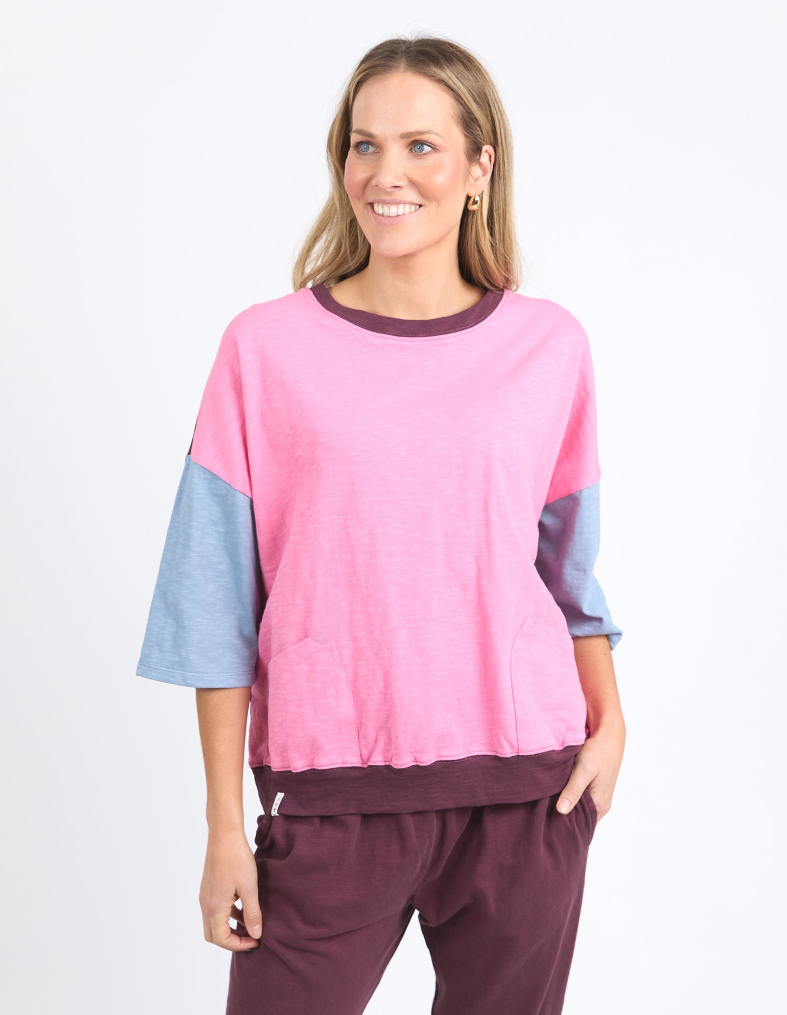 Image of A woman modeling a Elm color-blocked knit top with a heather pink body, light blue three-quarter sleeves, and dark plum trim, paired with matching dark plum pants against a plain white studio background.