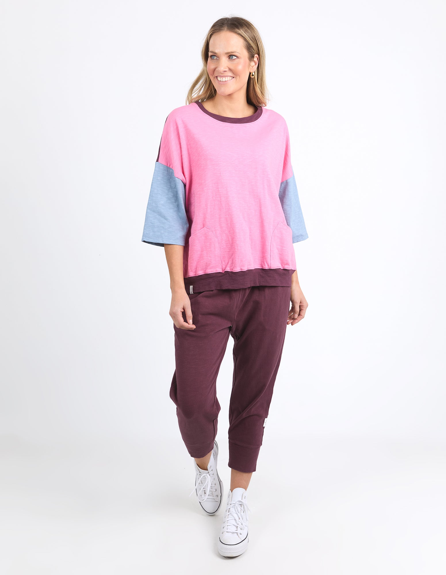 Image of A woman modeling a Elm color-blocked knit top and matching jogger pants set. The top features a bright pink body, light blue three-quarter sleeves, and a dark plum crew neck and hem.