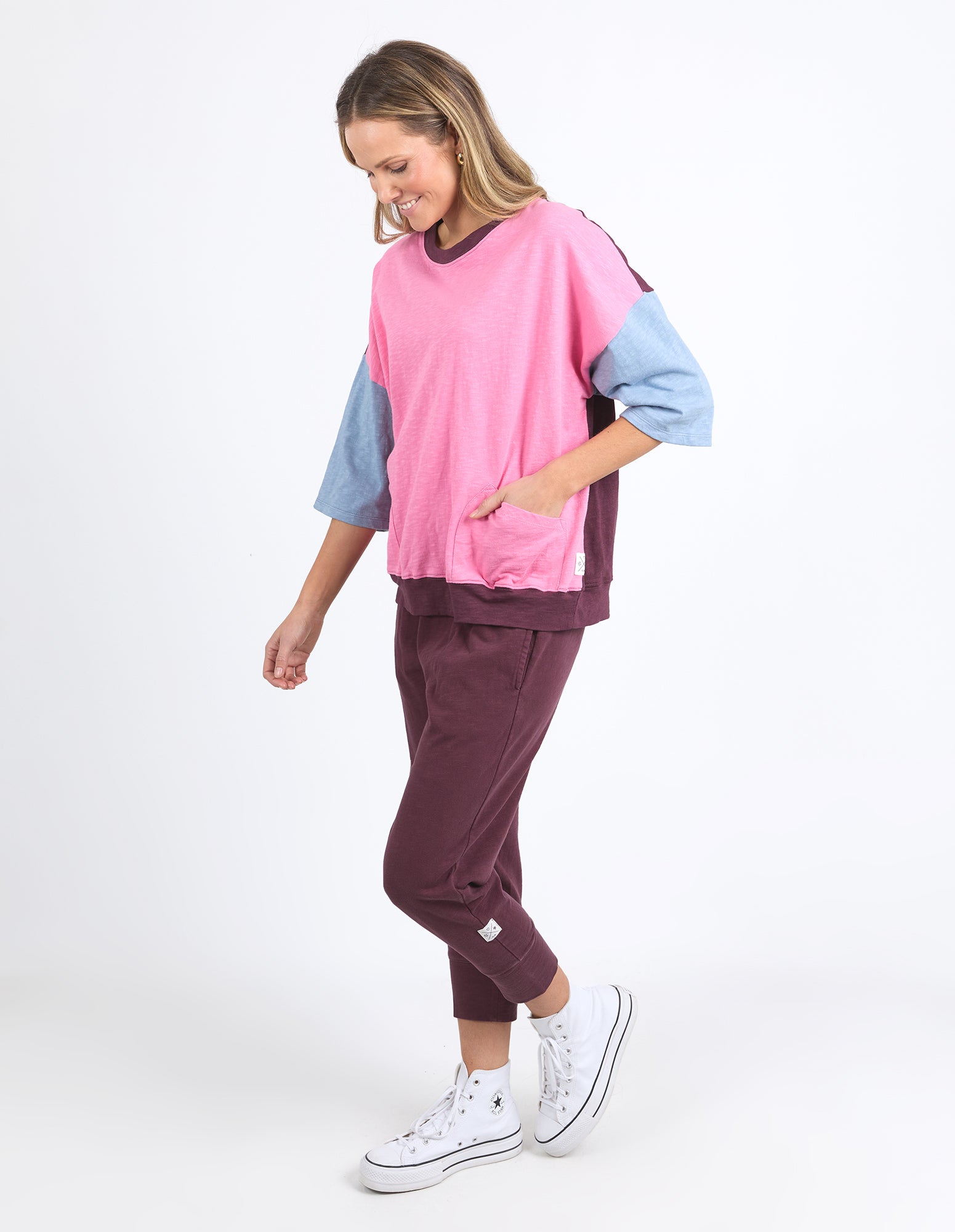 Image of A woman modeling a Elm two-piece colorblock tracksuit with a pink, light blue, and dark purple sweatshirt and matching joggers, paired with white sneakers, against a plain white studio background.
