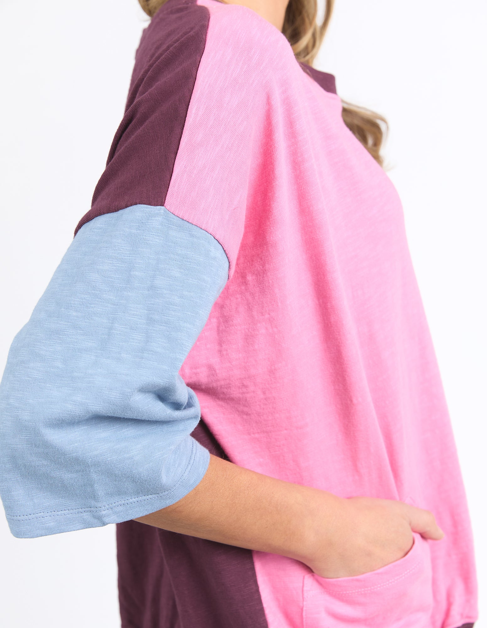 A close-up side view of a woman wearing a Elm colorblock raglan t-shirt with a pink body, dark purple shoulder yoke, and light blue heathered three-quarter sleeves.