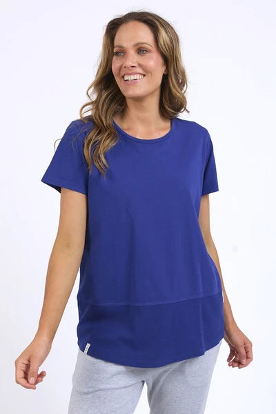 A woman modeling a Elm royal blue short-sleeve tunic top paired with light gray jogger pants and beige sneakers, standing against a plain white studio background.