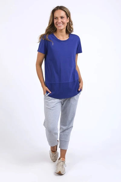 A woman modeling a Elm royal blue short-sleeve tunic top paired with light gray jogger pants and beige sneakers, standing against a plain white studio background.