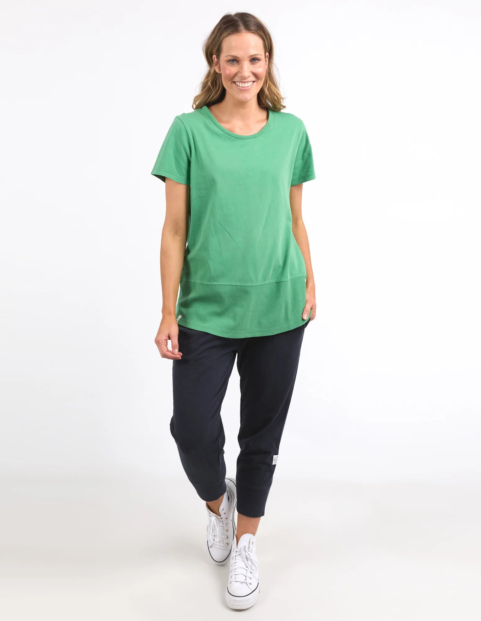 Image of A woman modeling a Elm green short-sleeve crewneck t-shirt paired with dark navy pants and white sneakers, standing against a plain white studio background.