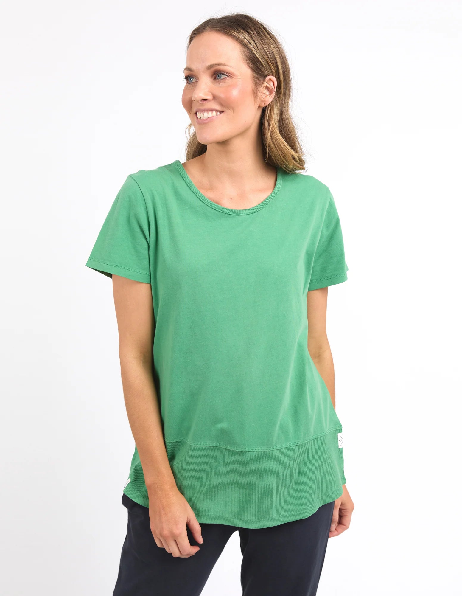 Image of A woman wearing a Elm green short-sleeve crewneck top with a layered hem and dark pants, standing against a plain white studio background.