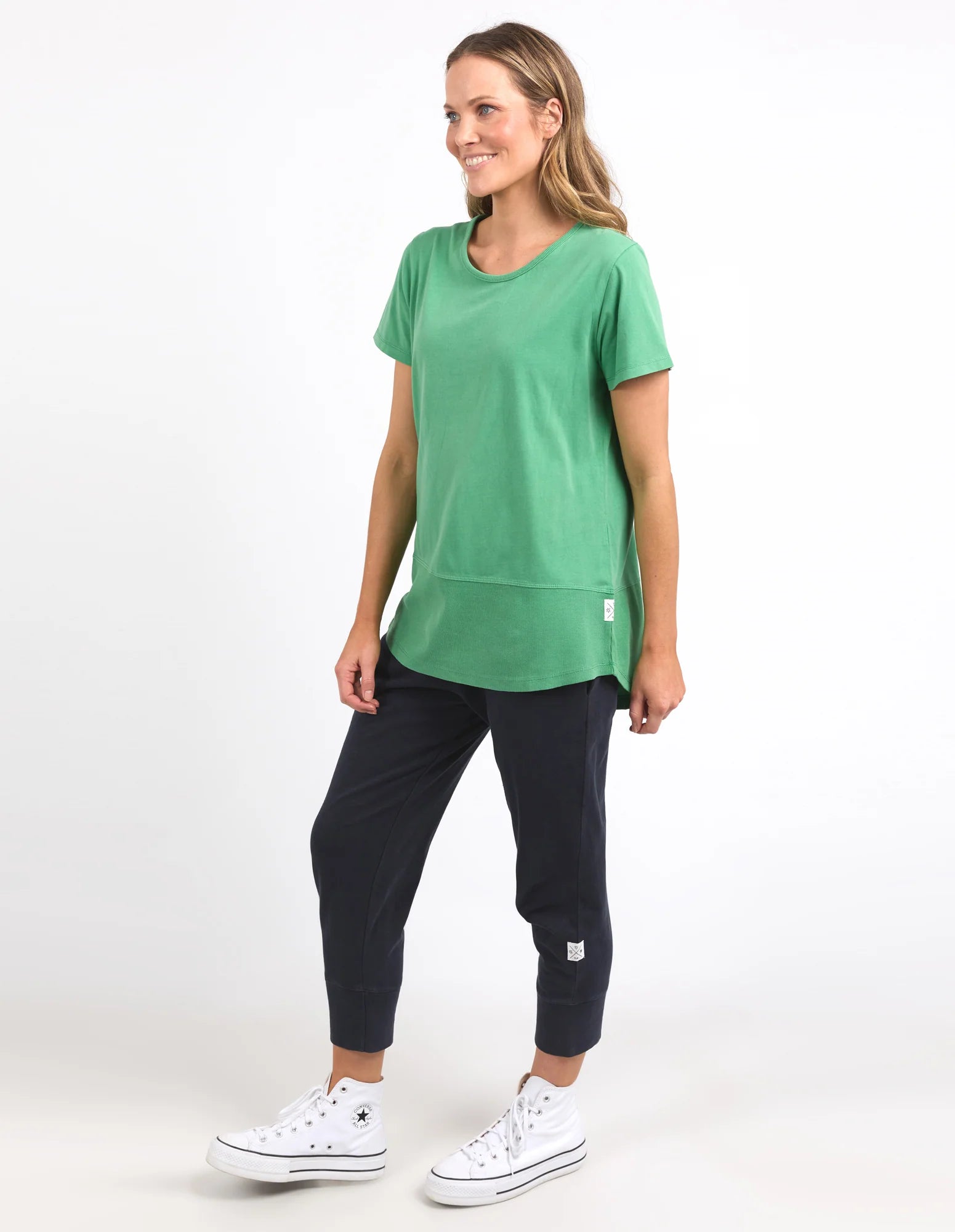 Image of A woman modeling a Elm green short-sleeve tunic top with a front pocket, paired with dark cropped pants and white sneakers, against a plain white studio background.