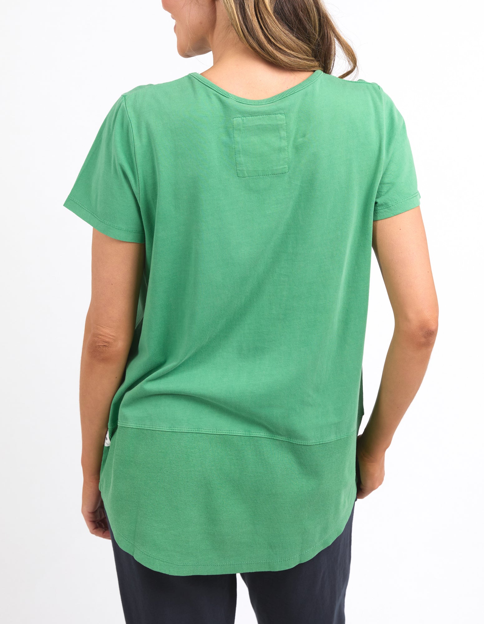 Image of A woman shown from the back wearing a Elm green short-sleeve top with a curved hem and a small rectangular patch pocket on the upper back, paired with dark pants against a plain white background.