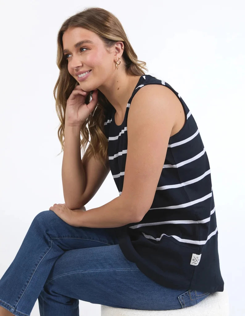 A woman wearing a Elm navy blue sleeveless tank top with white horizontal stripes and blue jeans, sitting against a plain white studio background.