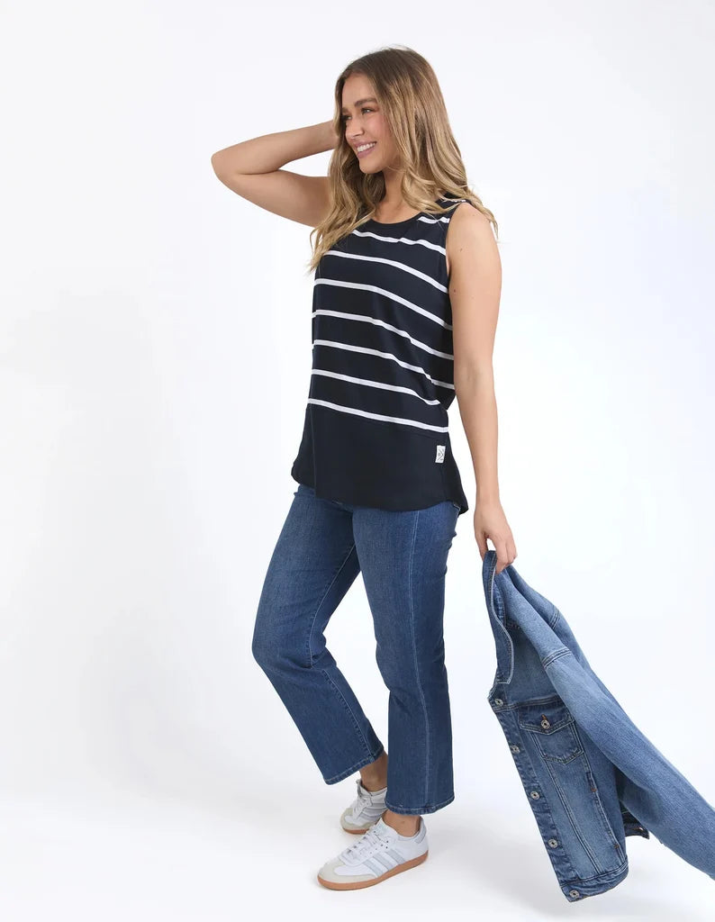 A woman modeling a Elm sleeveless black tank top with thin white horizontal stripes, paired with blue jeans and white sneakers, holding a light blue denim jacket against a plain white studio background.