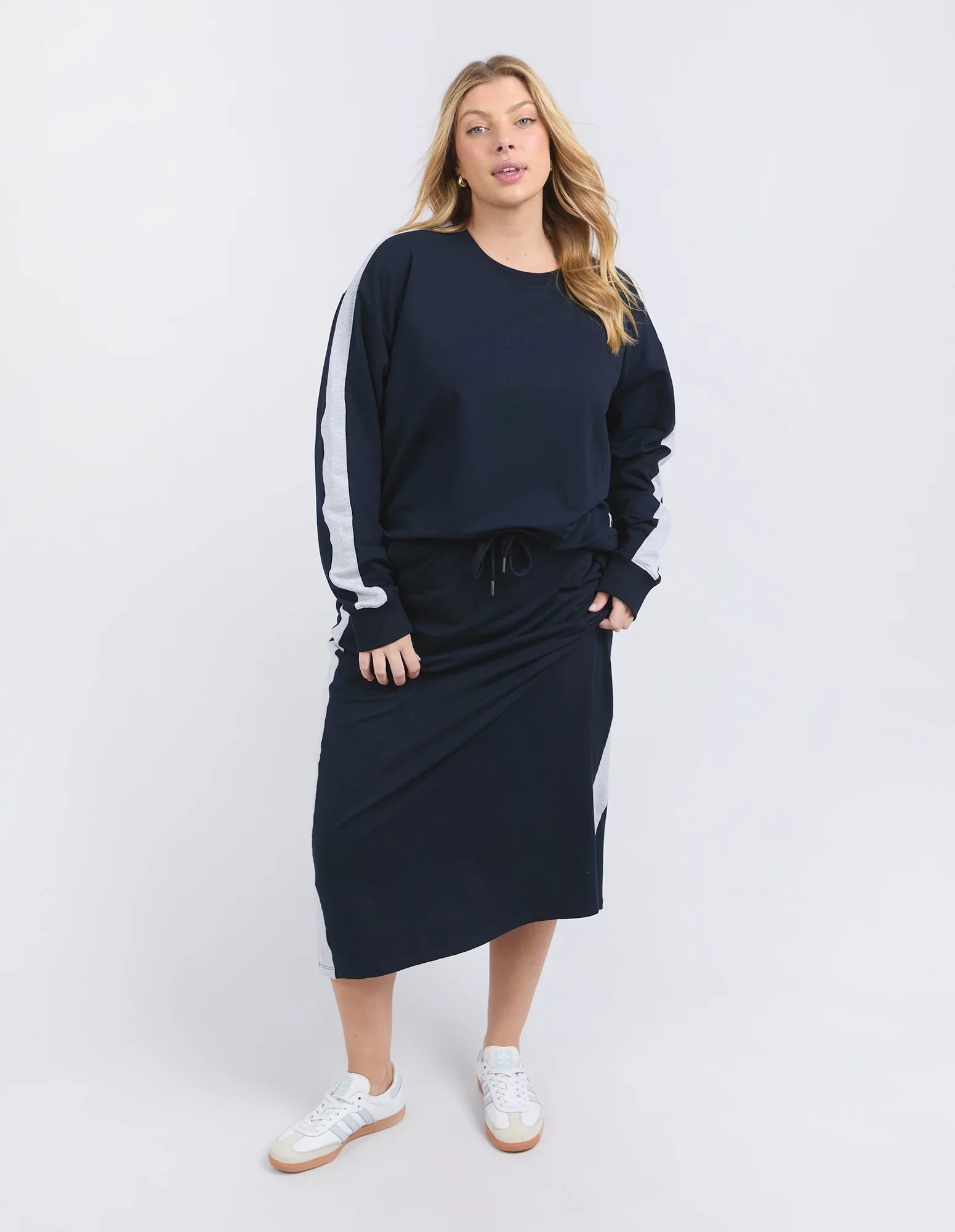 Image of A woman modeling a Elm navy blue lounge set consisting of a long-sleeve sweatshirt with white side stripes and a matching midi skirt, paired with white sneakers, against a light gray studio background.