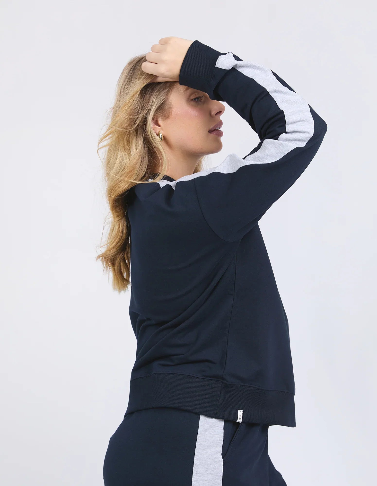 Image of A woman wearing a Elm navy blue athletic jumper with white stripe details on the sleeves and matching navy pants, posing against a light gray studio background.