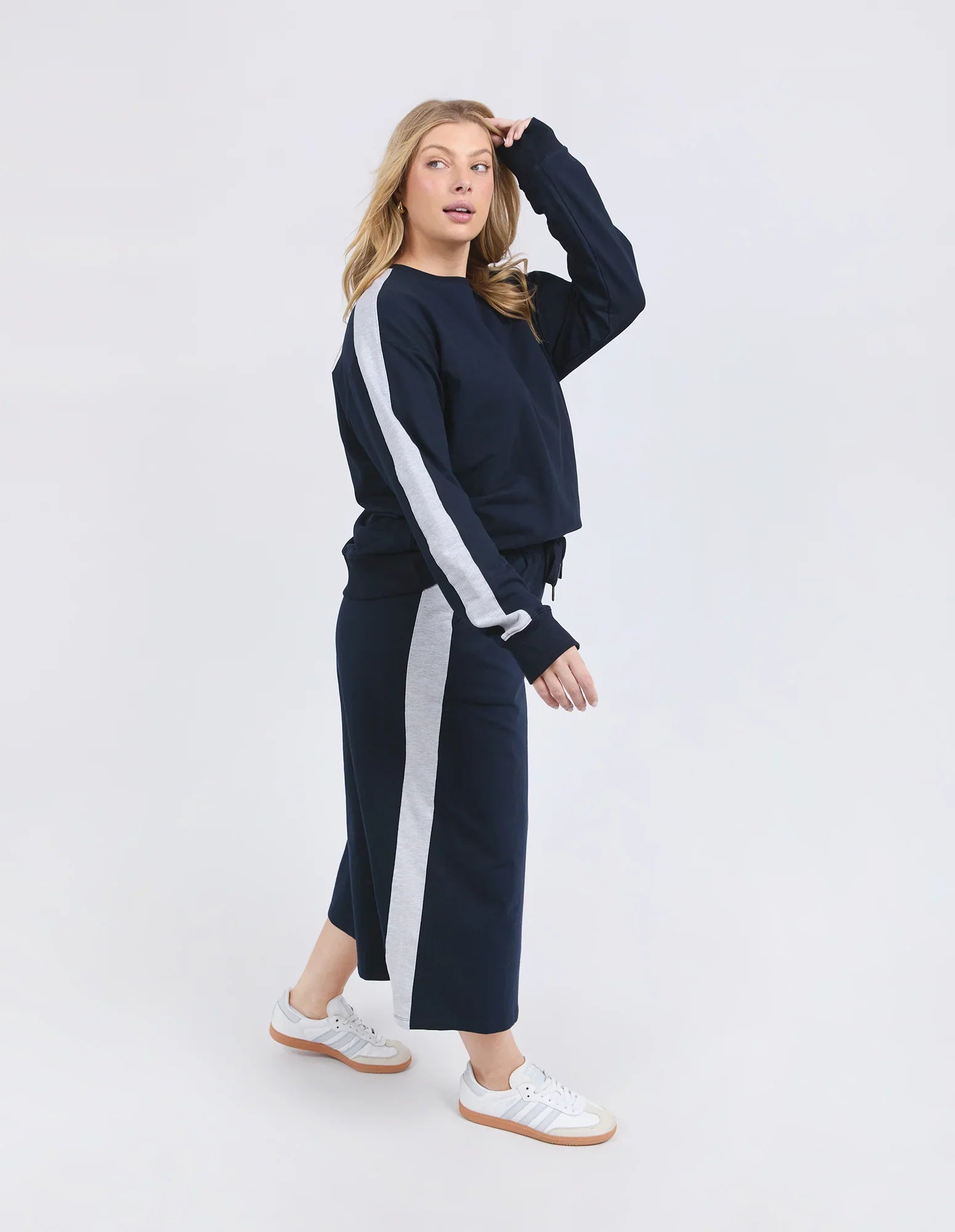 A woman modeling a matching Elm navy blue sweatshirt and cropped wide-leg trousers set, both featuring a white vertical stripe down the side, paired with white sneakers, against a plain light gray studio background.