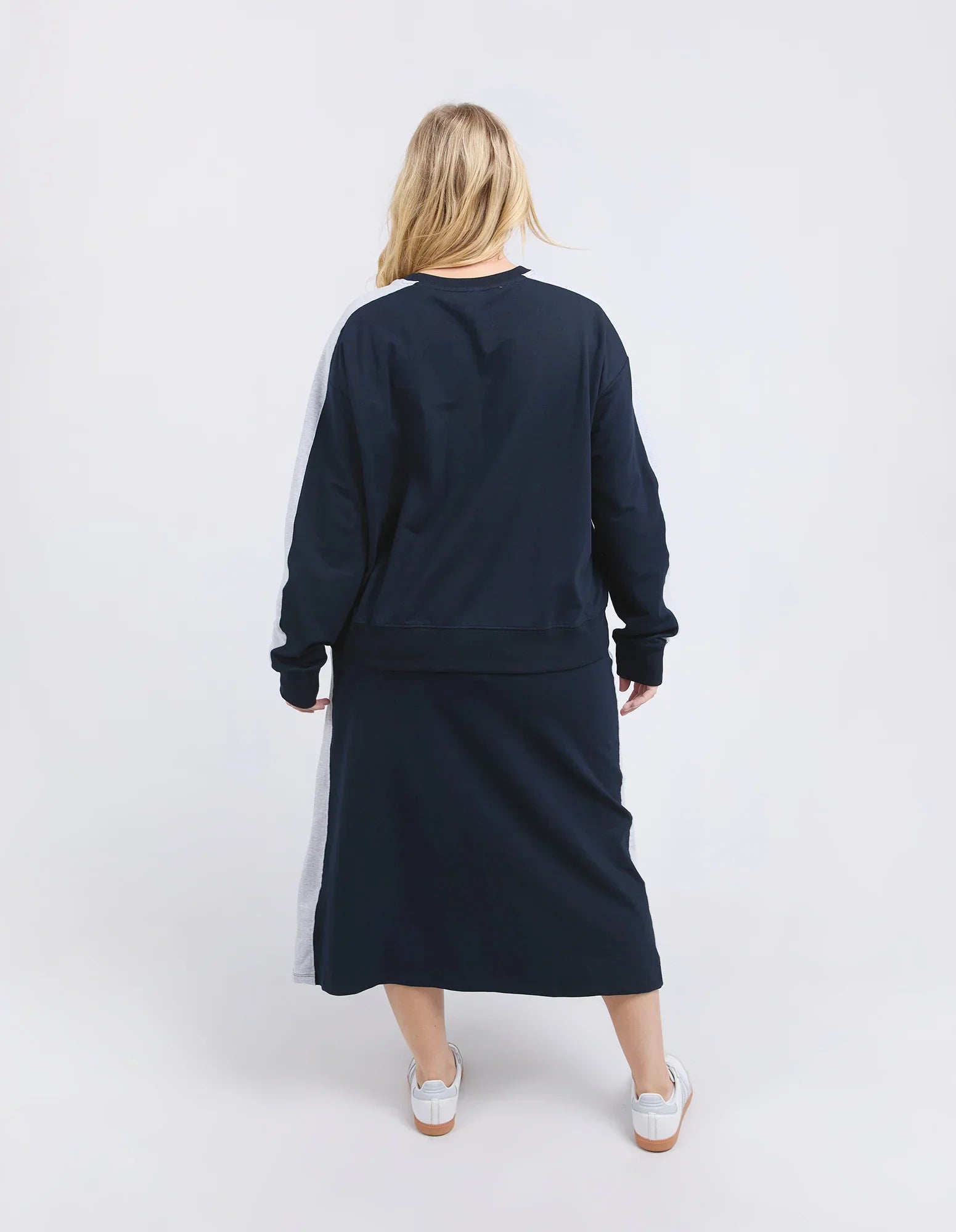 Image of A woman viewed from behind wearing a Elm navy blue long-sleeve sweatshirt and matching midi skirt set with light gray side stripes, paired with white sneakers, against a plain light gray studio background.