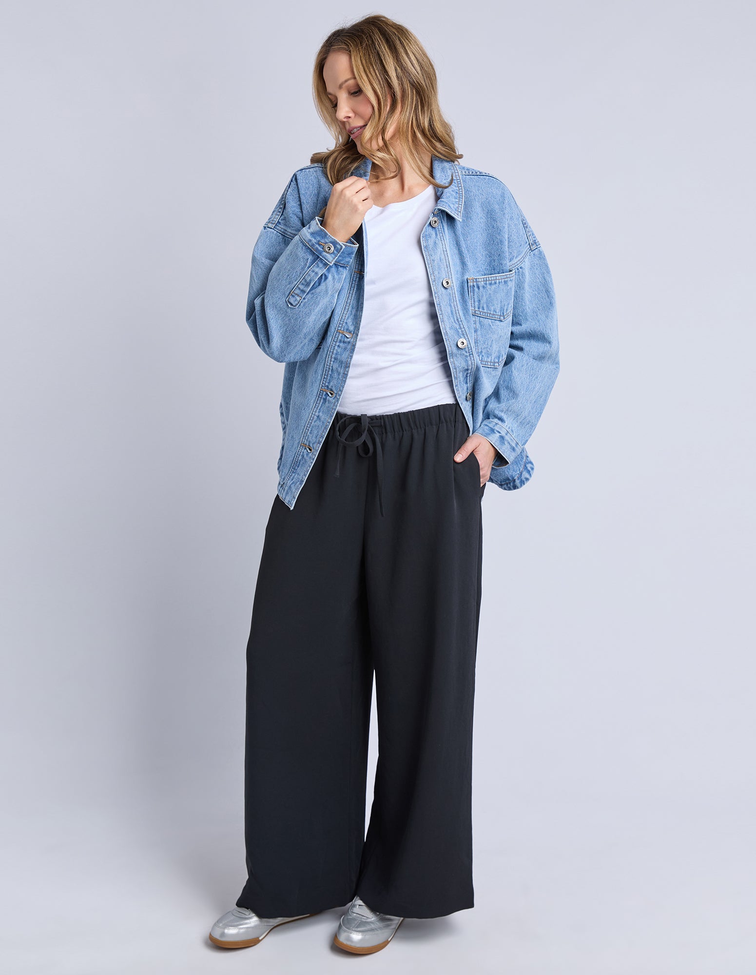 Image of Woman wearing a denim jacket, white shirt, and Elm Sophie black wide-leg pants on a light grey background