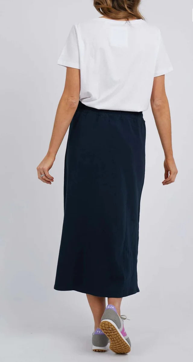 Image of A woman shown from the back wearing a white short-sleeve t-shirt and a Elm dark navy midi skirt, paired with gray sneakers, against a light gray studio background.