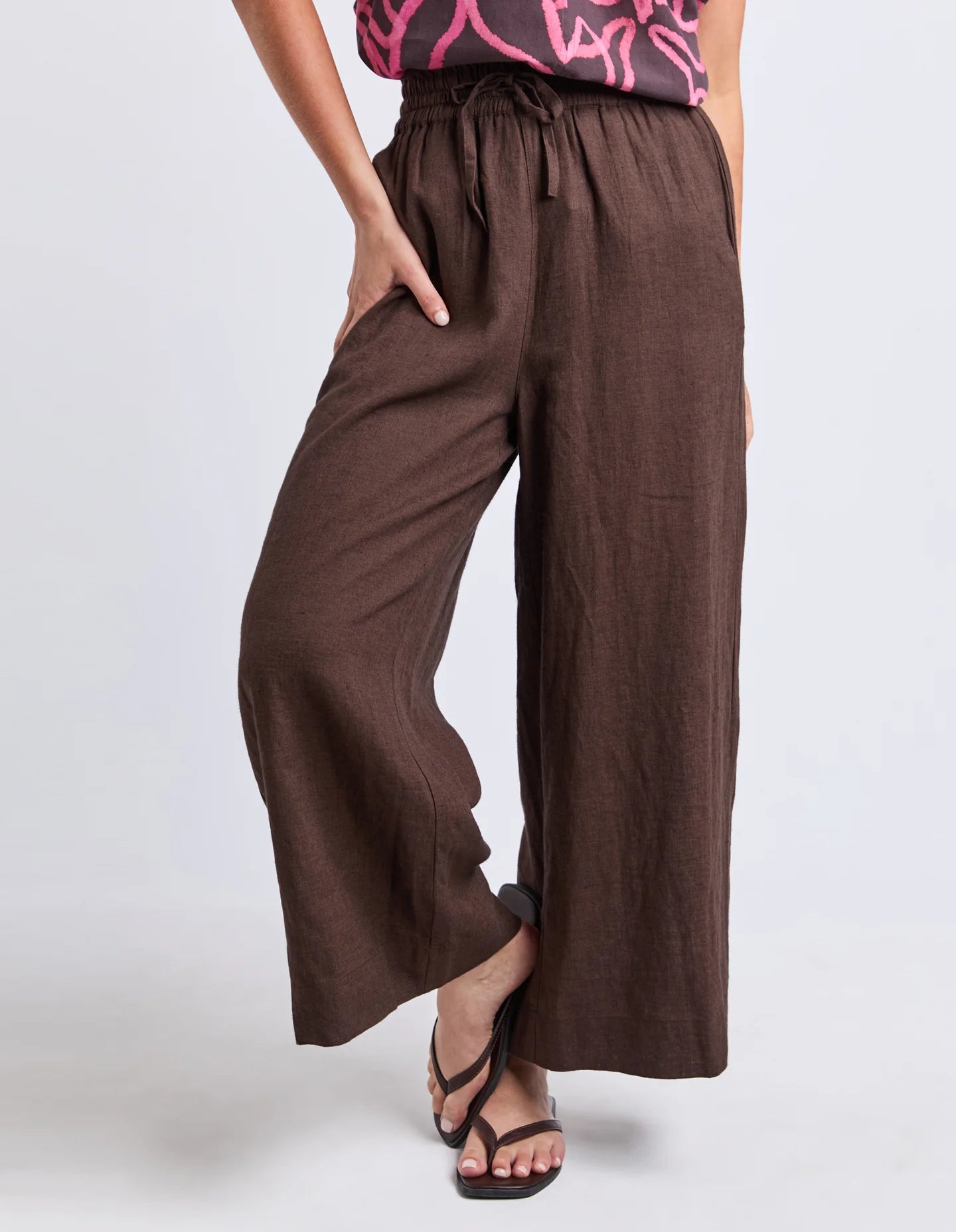 Image of Woman wearing Elm chocolate brown wide leg pants paired with a printed blouse on a plain background 