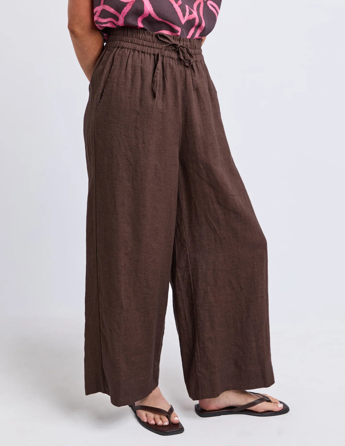 Woman wearing Elm chocolate brown wide leg pants paired with a printed blouse on a plain background 