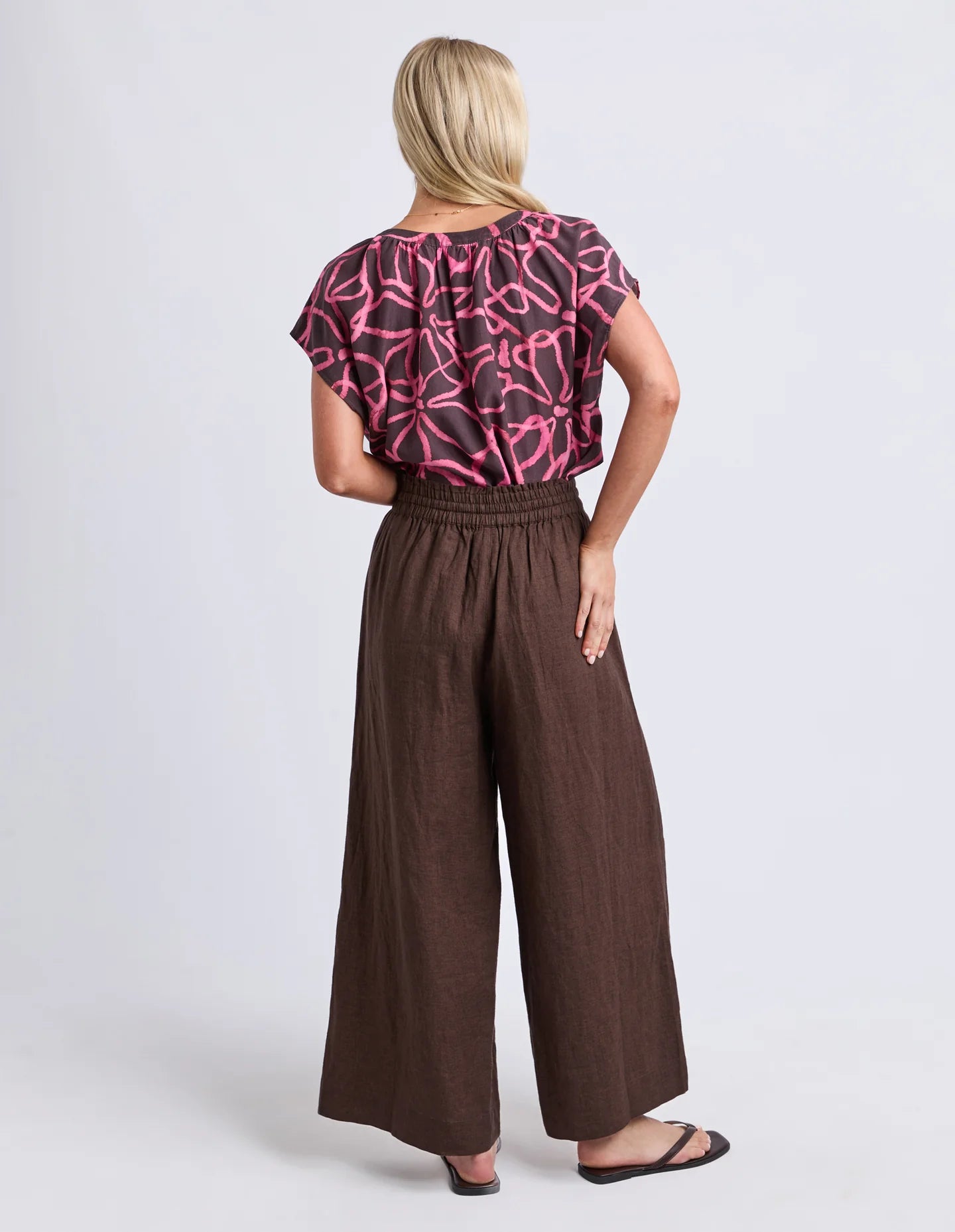 Image of Woman wearing Elm chocolate brown wide leg pants paired with a printed blouse on a plain background 