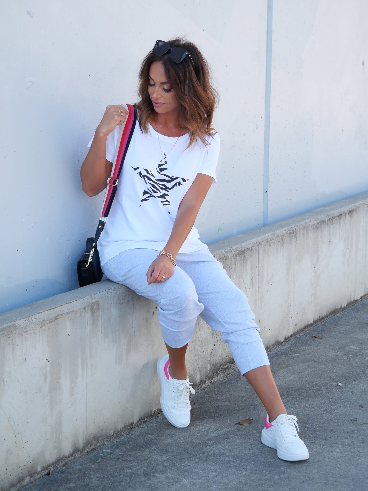 Image of A woman sitting on a concrete ledge against a light blue wall, wearing a white Elm graphic t-shirt, white cropped pants, and white sneakers with pink accents. She is carrying a black shoulder bag with a red and black strap.