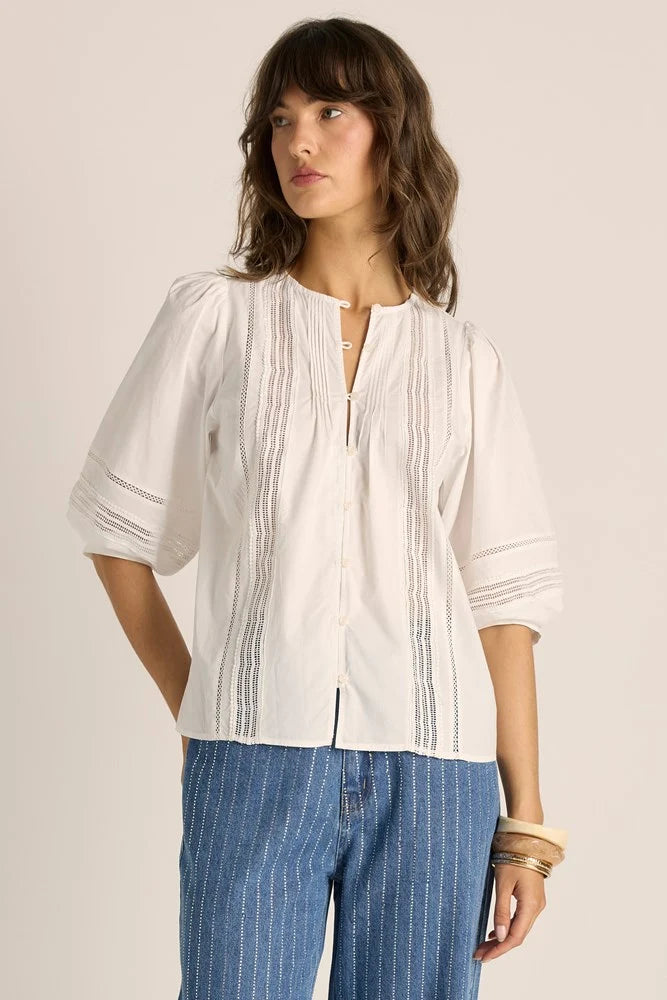 Image of A woman wearing a Fate & Becker white short-sleeve blouse with vertical embroidered stripes and a split neckline, paired with blue pinstriped pants, against a plain beige studio background.