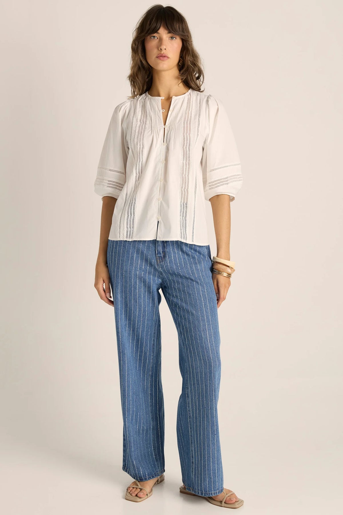 A woman modeling a Fate & Becker white short-sleeve blouse with vertical pleats and a split neckline, paired with blue pinstriped wide-leg pants and beige sandals, against a plain light beige studio background. 
