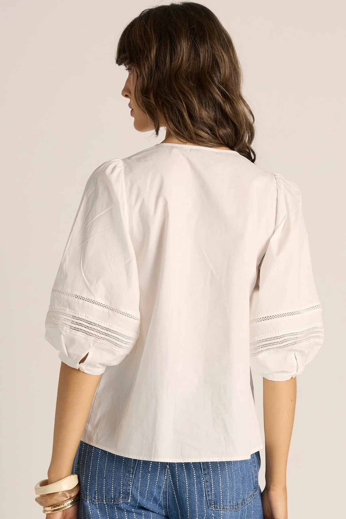 A woman shown from the back wearing a Fate & Becker white short-sleeve blouse with puffed sleeves and delicate horizontal embroidery near the cuffs, paired with blue jeans against a light beige studio background.